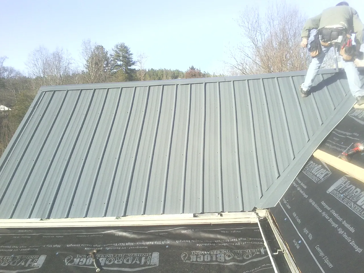 Crew installing dark green standing seam panels over synthetic underlayment in Elliston