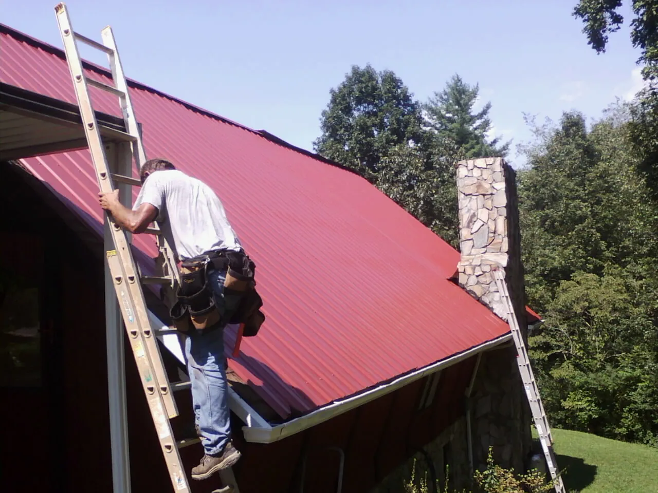 Roofer on a ladder beside a red metal roof and stone chimney — Soffit & Fascia Repair by Roofing Brothers in Elliston